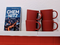 Vier rote Tassen auf einem Regal neben einem Buch über Chemnitz.